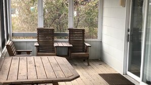 Una terraza de madera con una mesa y dos sillas, ventanas amplias y un gran ventanal corredizo.