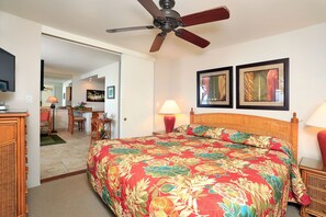 Une chambre d’hôtel comprenant un lit, un ventilateur de plafond, deux tableaux encadrés, un bureau avec une chaise et un téléviseur.