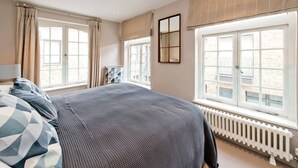 Une chambre avec un grand lit, deux fenêtres avec des rideaux, un radiateur et une vue sur l’extérieur.