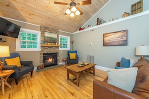 A cozy living room with a fireplace, wooden flooring, and a ceiling fan.