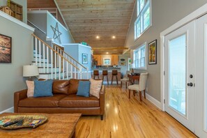 A spacious living area with a brown leather sofa, wooden flooring, a staircase, and a kitchen in the background.
