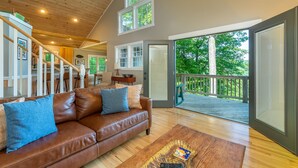 A spacious living room with a brown leather sofa, wooden flooring, and a large sliding glass door leading to a balcony with a view of trees. - Sunset Mtn Lodge! Hot tub+Fire pit, Fireplace, Hiking, Private, Close to Town! (Spruce Pine)
