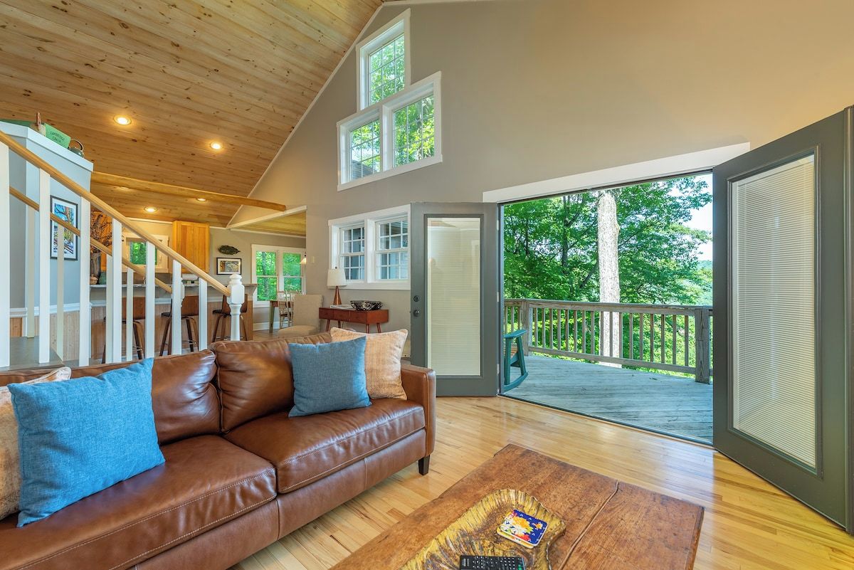 A spacious living room with a brown leather sofa, a wooden coffee table, and a view of a deck through large windows.