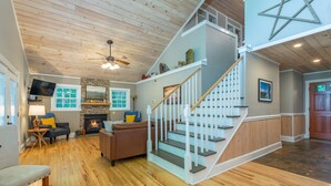 A spacious living area with a fireplace, a staircase, and a television. - Sunset Mtn Lodge! Hot tub+Fire pit, Fireplace, Hiking, Private, Close to Town! (Spruce Pine)