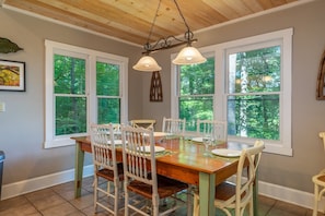 A dining area with a wooden table and chairs, large windows with a view of trees, and a hanging light fixture. - Hot Tub+Fire Pit, Mtn Sunsets, Fireplace & Grill! (Spruce Pine)