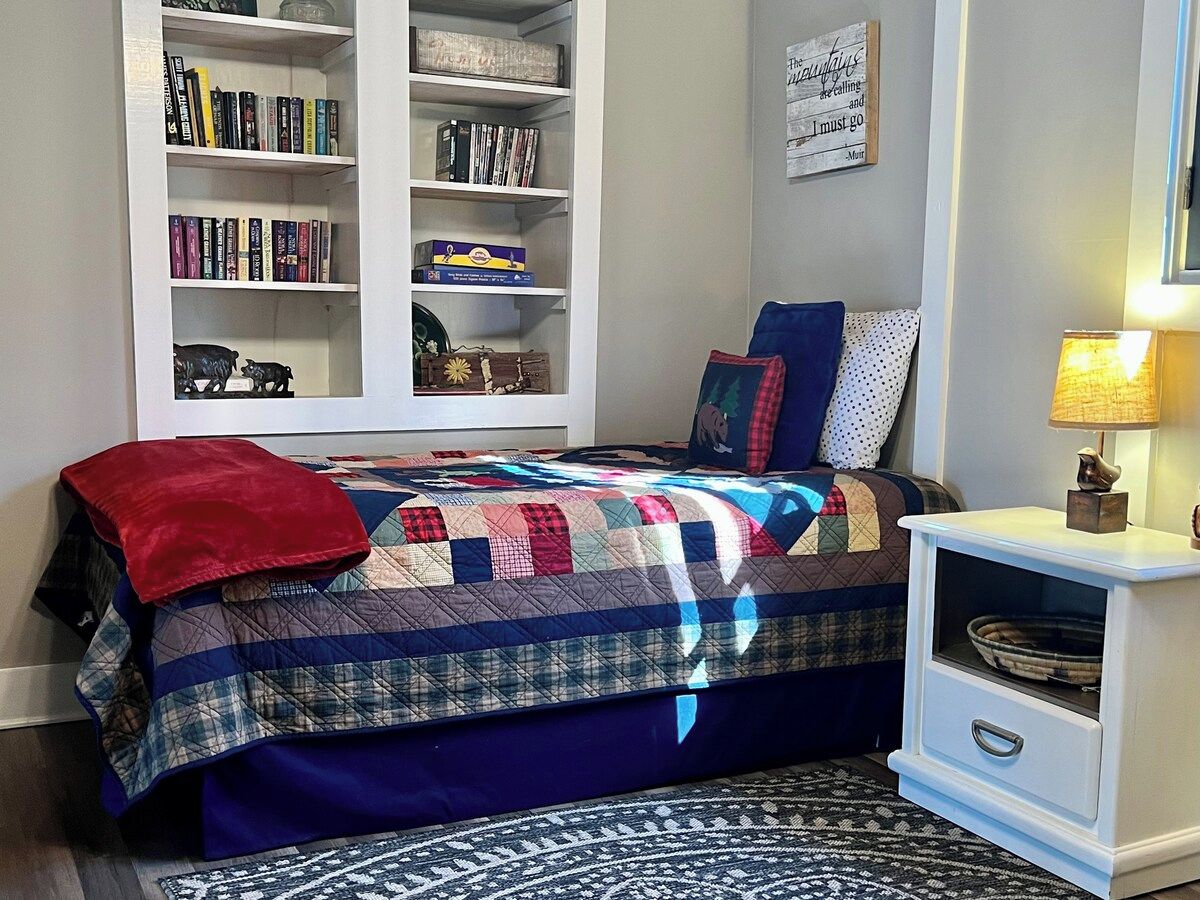 A bedroom with a bed, a bookshelf, a desk, and a wall with a quote.