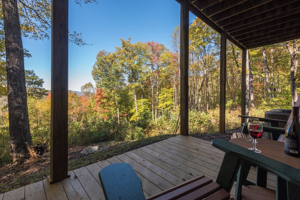 A wooden deck with a table and chairs, overlooking a forested area with trees and a clear sky.