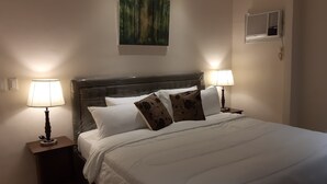 A neatly made bed with white bedding and floral pillows, two bedside lamps, a wall-mounted air conditioner, and a painting of a forest. - Affordable Luxury 2BR Condo in Davao City (Davao City)