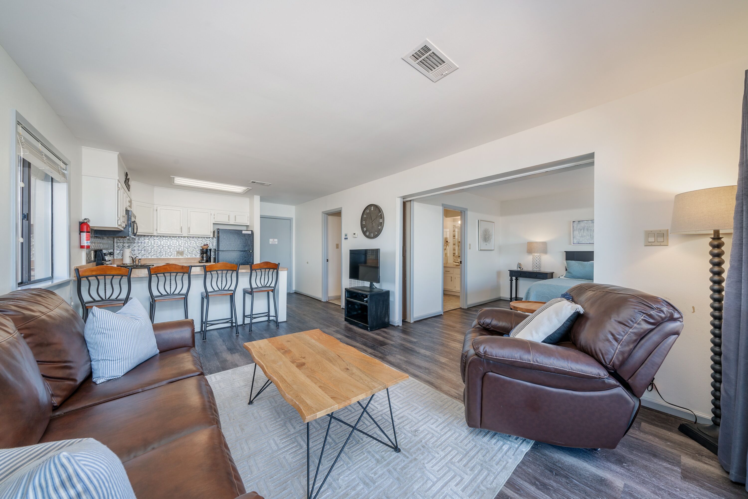 A modern living room with a sofa, a wooden coffee table, and a leather armchair. There is a kitchen area with a dining set and a TV.