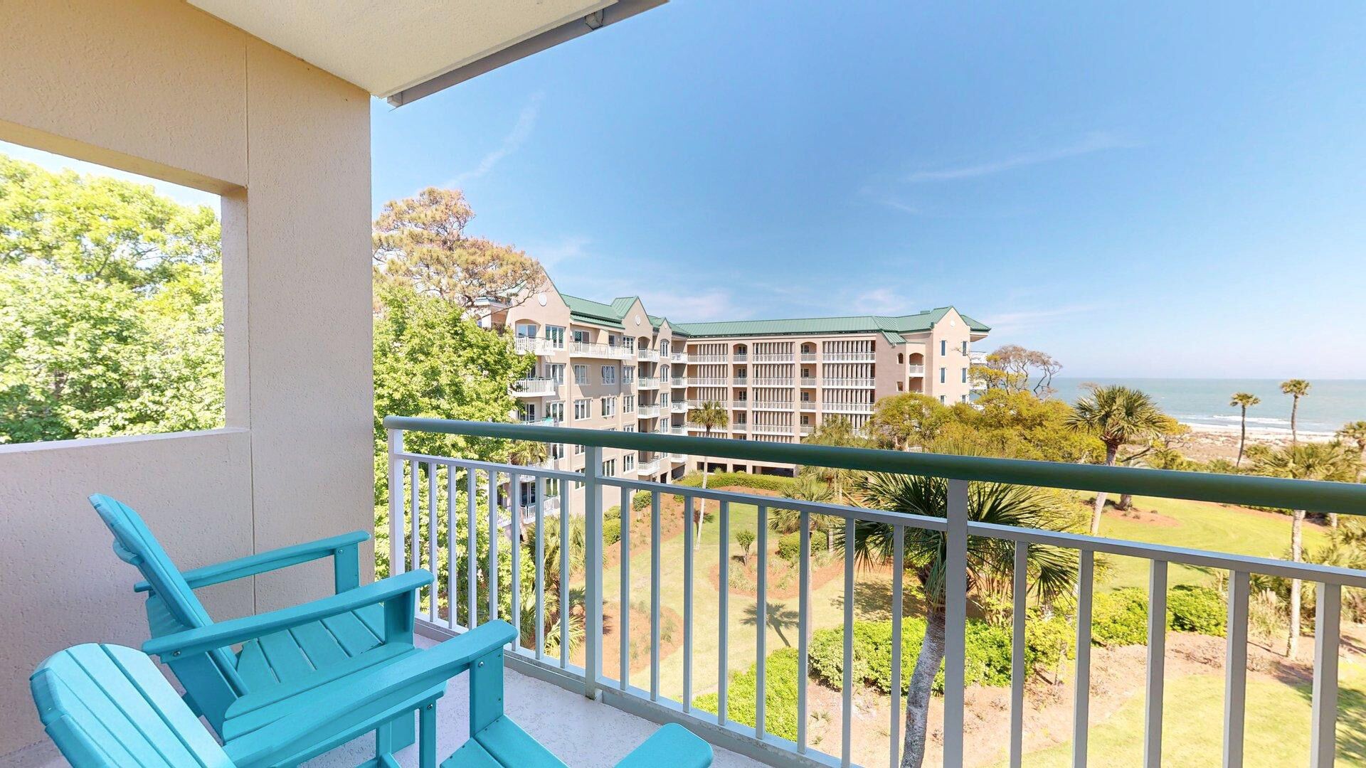 Ein Balkon mit türkisfarbenen Stühlen, Blick auf ein Hotel mit mehreren Etagen und einem begrünten Dach, sowie ein Strand im Hintergrund.