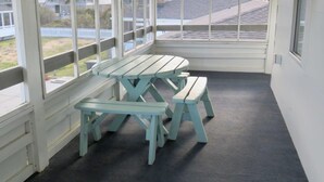 A covered outdoor seating area with a picnic table and benches, large windows, and a view of buildings and a fence. - Saltaire Cottages 6: 2BR Steps to Private Beach Access Screen Porch Pool (Kitty Hawk)