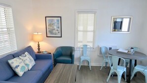 A living room with a blue sofa, a coffee table, and a dining area with a table and chairs. - Saltaire Cottages 5: 2BR Steps to Private Beach Access Screen Porch Pool (Kitty Hawk)