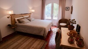 A bedroom with a bed, bedside lamps, a wooden headboard, a window with curtains, a wicker chair, a small table with a plant, and a woven rug. - Guest Room in nature in Minorca : Adults Only (Ferreries)