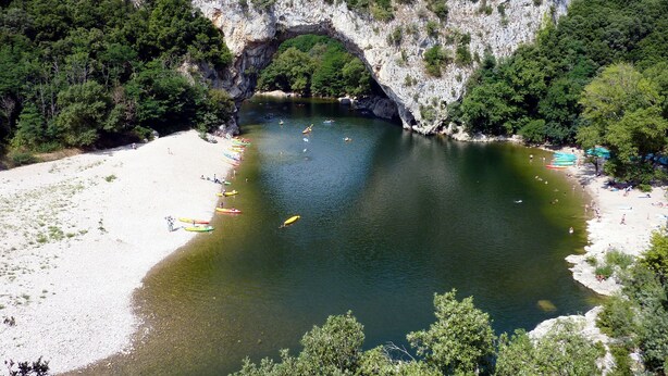 A natural arch formation over a river with a sandy beach and boats.