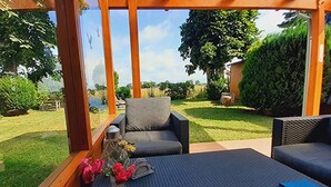 A covered outdoor seating area with two sofas and a table, surrounded by greenery and a view of a lake. - House Dina Ahrenshoop (Ahrenshoop)