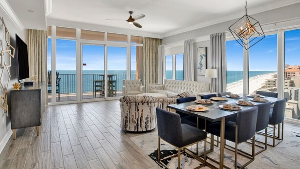 A spacious living room with a large sofa, a dining area with a table set for four, and a balcony with a view of the ocean. - Phoenix Orange Beach 1201 | Orange Beach, AL (Orange Beach)