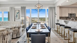 A modern living room with a dining area, large windows with a city view, a kitchen with a microwave, and a bar area with stools. - Phoenix Orange Beach 1201 | Orange Beach, AL (Orange Beach)