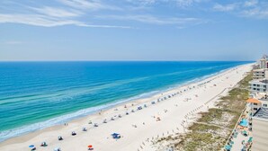 A beach with a long stretch of sand, umbrellas, and a building with balconies overlooking the ocean. - Phoenix Orange Beach 1201 | Orange Beach, AL (Orange Beach)