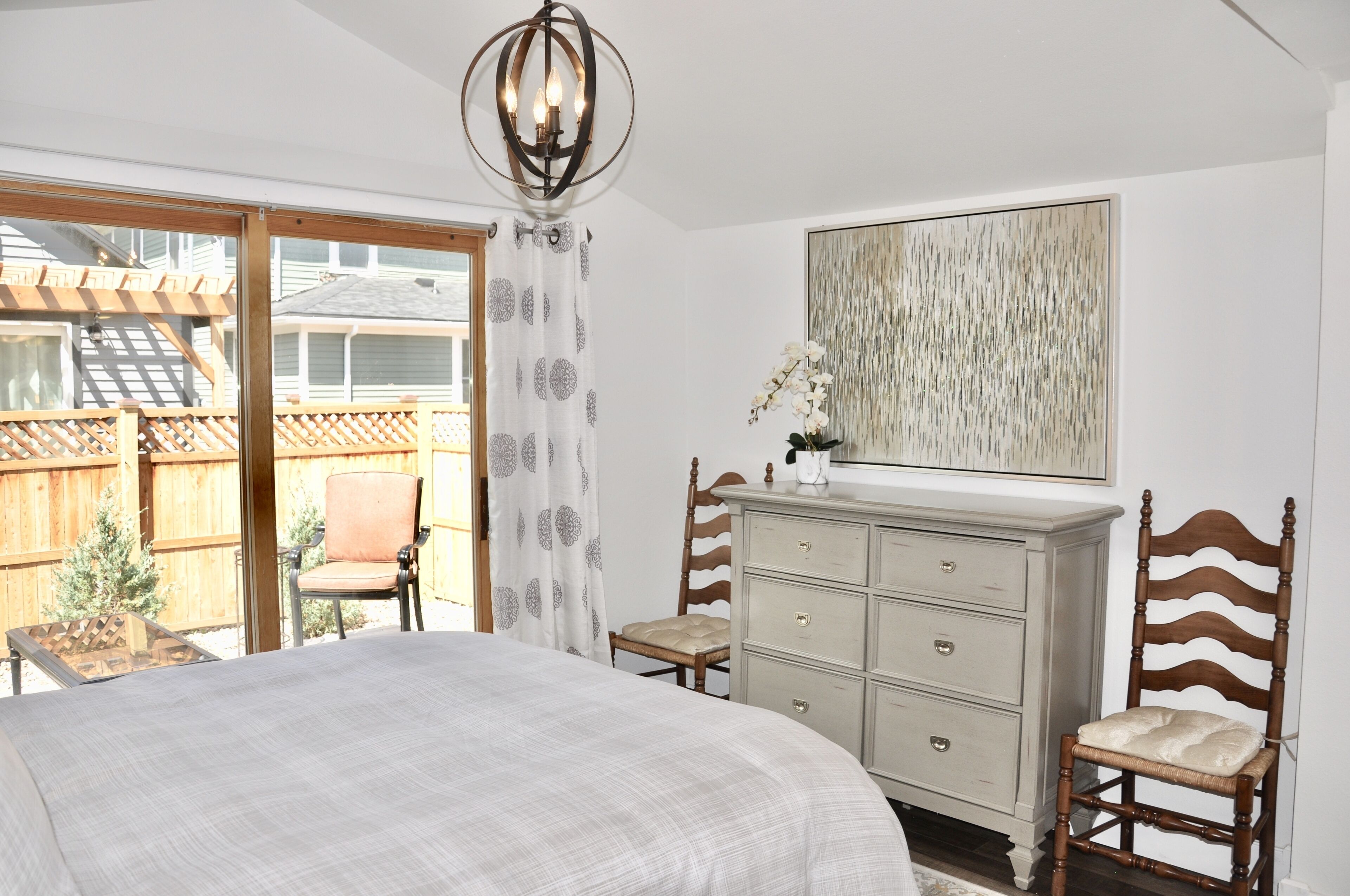 Une chambre avec un grand lit, une commode, deux chaises, un lustre et une vue sur une terrasse en bois.