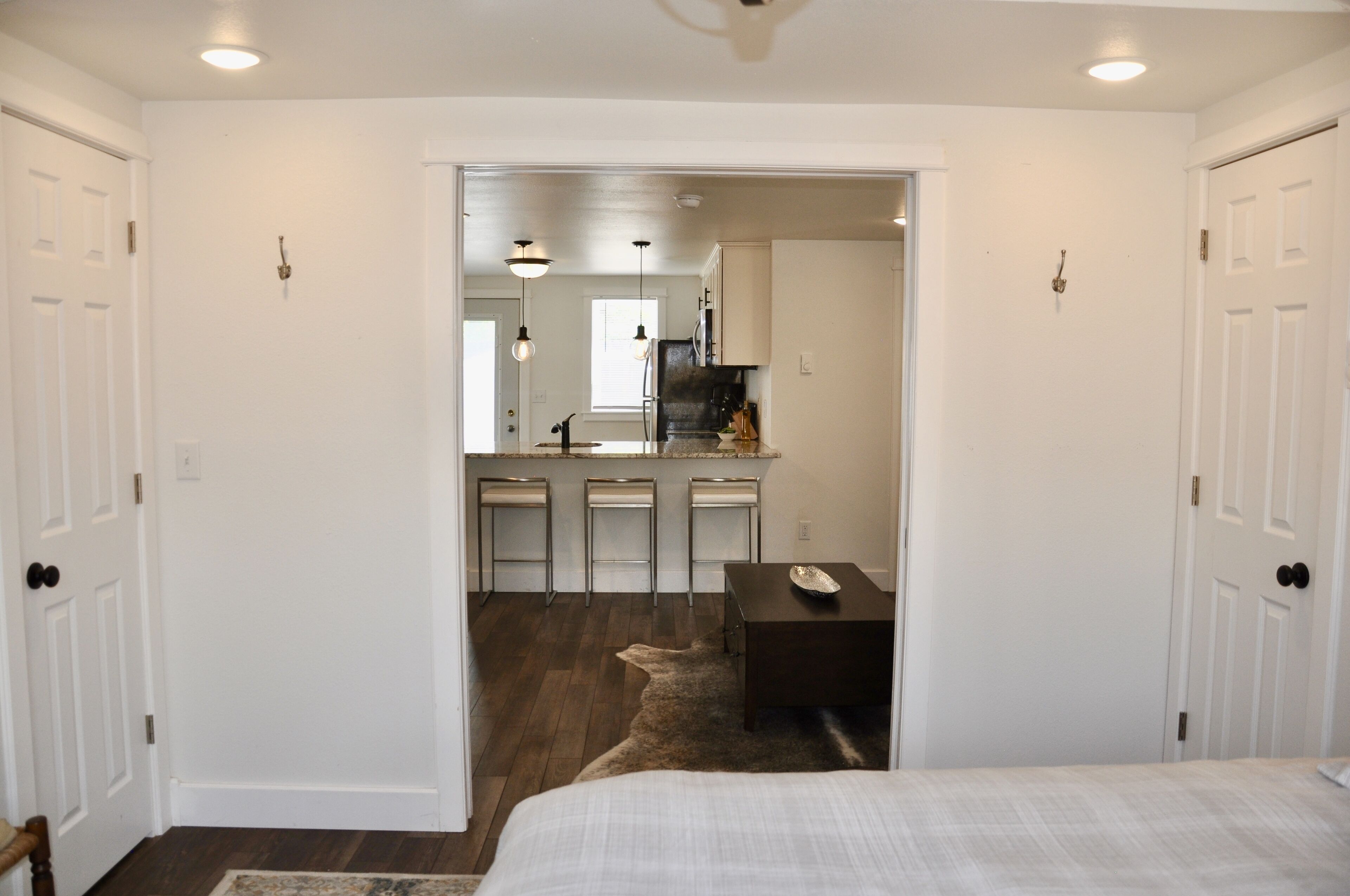 A bedroom with a large bed, a small desk, and a doorway leading to a kitchen area with a sink and bar stools.