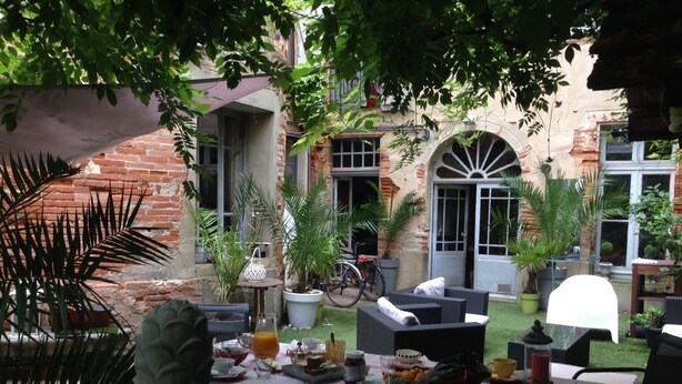 Un área de comedor al aire libre con una mesa dispuesta para una comida, rodeada de plantas en macetas y un edificio de ladrillo al fondo.