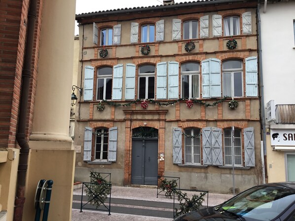 Un edificio de varios pisos con arquitectura tradicional, que presenta una entrada central con una puerta oscura, numerosas ventanas con contraventanas y decoraciones navideñas.