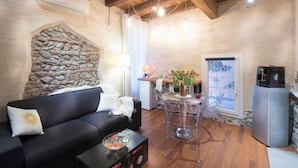 A cozy living room with a stone wall, a black sofa, a clear glass table, and a kitchen area with a coffee machine. - Abbraccio di Romeo e Giulietta (Verona)