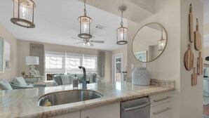 A modern kitchen with a marble countertop, stainless steel sink, and a dishwasher. There are hanging lights, a ceiling fan, and a large mirror on the wall. - ☀️Beachfront Complex-Top Floor-30A-Driftwood Cottage (Santa Rosa Beach)