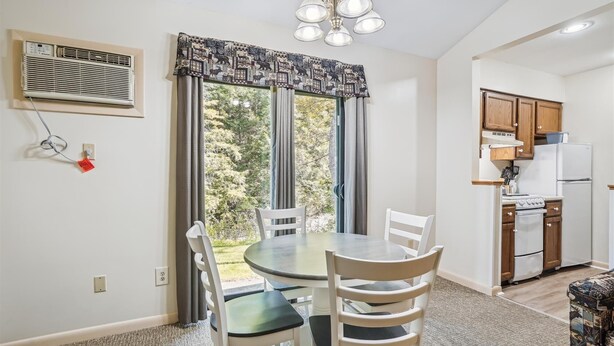 A dining area with a round table and chairs, a window with curtains, a kitchen with wooden cabinets and appliances, and a ceiling light fixture.