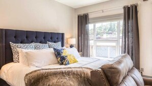 A bedroom with a tufted headboard, a large window with curtains, and a view of the outdoors.