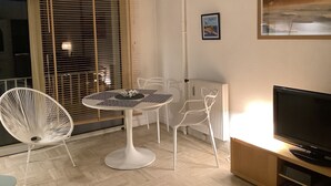 Un salon moderne avec une table ronde blanche, deux chaises blanches, un téléviseur noir posé sur un meuble en bois, un radiateur et un tableau encadré accroché au mur.