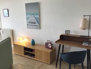 Ein modernes Wohnzimmer mit einem Holz-TV-Schrank, einem Vintage-Radio, einer Lampe, einem Bilderrahmen mit einem Blick auf einen Pier und einer kleinen Uhr auf einem Regal.