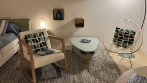 Une pièce chaleureuse avec une chaise en bois, une petite table et une lampe sur pied. On y trouve un lit avec des oreillers et un tableau encadré accroché au mur.