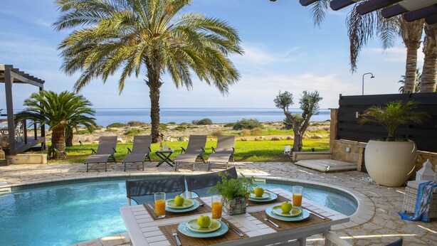 A poolside dining area with a table set for two, overlooking a beach with palm trees and a view of the ocean. - Protaras Seafront Villa El Greco (Protaras)