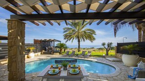 A poolside dining area with a table set for four, surrounded by lounge chairs, a stone pillar, and a view of the ocean. - Protaras Seafront Villa El Greco (Protaras)