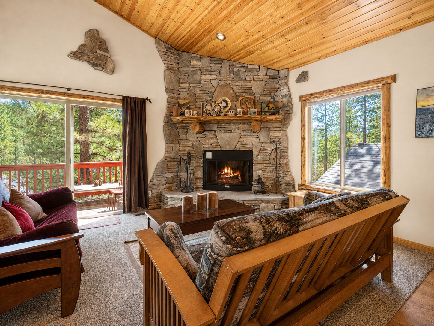 A cozy living room with a stone fireplace, wooden furniture, and large windows offering a view of the outdoors.