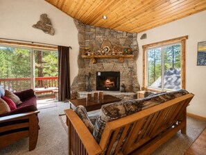 A cozy living room with a stone fireplace, wooden furniture, and large windows offering a view of the outdoors. - Monterey Pines | Mountain Views, Gas Fireplace & Walk to Alpine Zoo (Big Bear Lake)