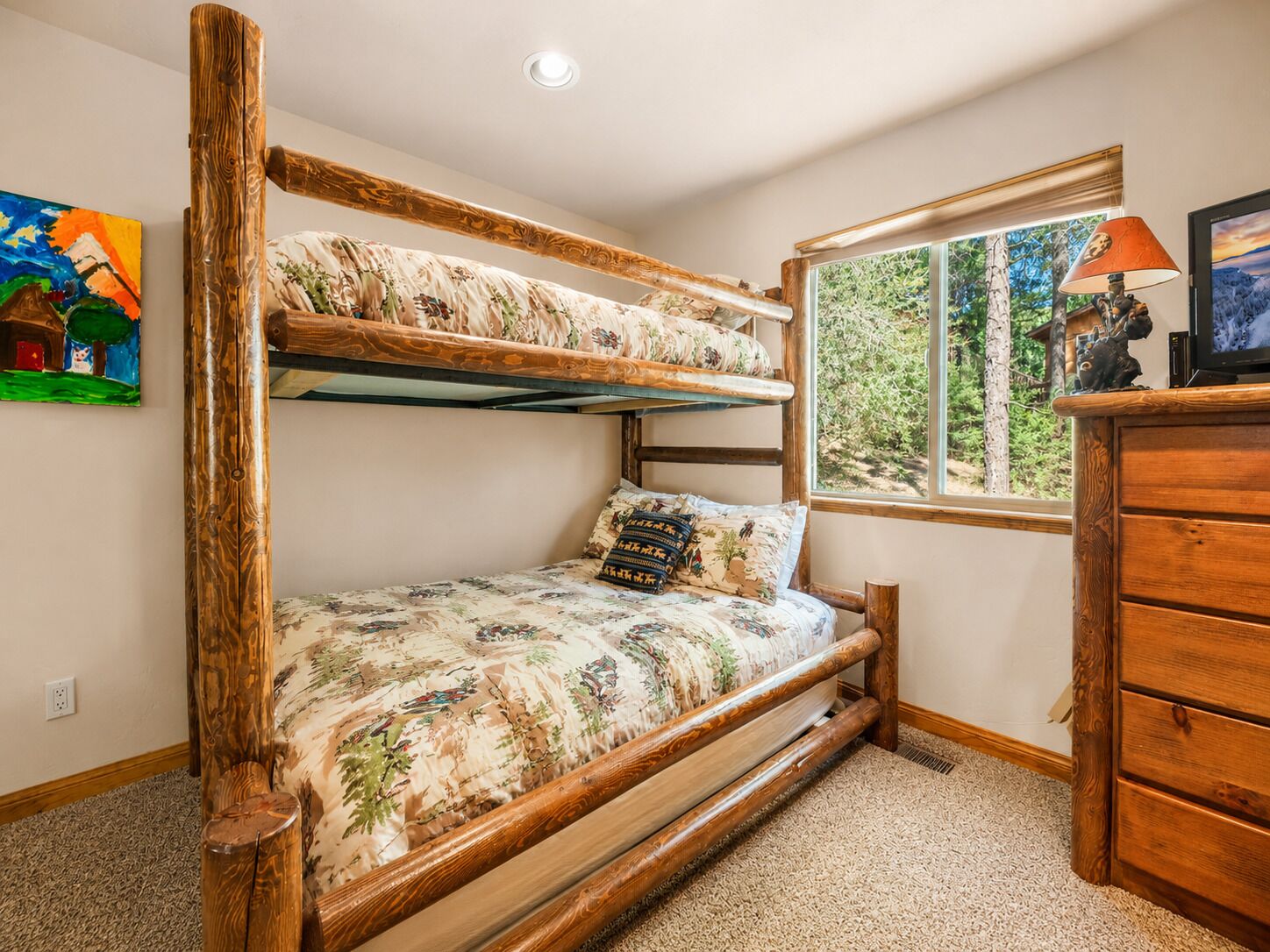Een stapelbed met houten frame en bloemenbeddengoed, een raam met uitzicht op bomen, en een houten dressoir met een lamp.