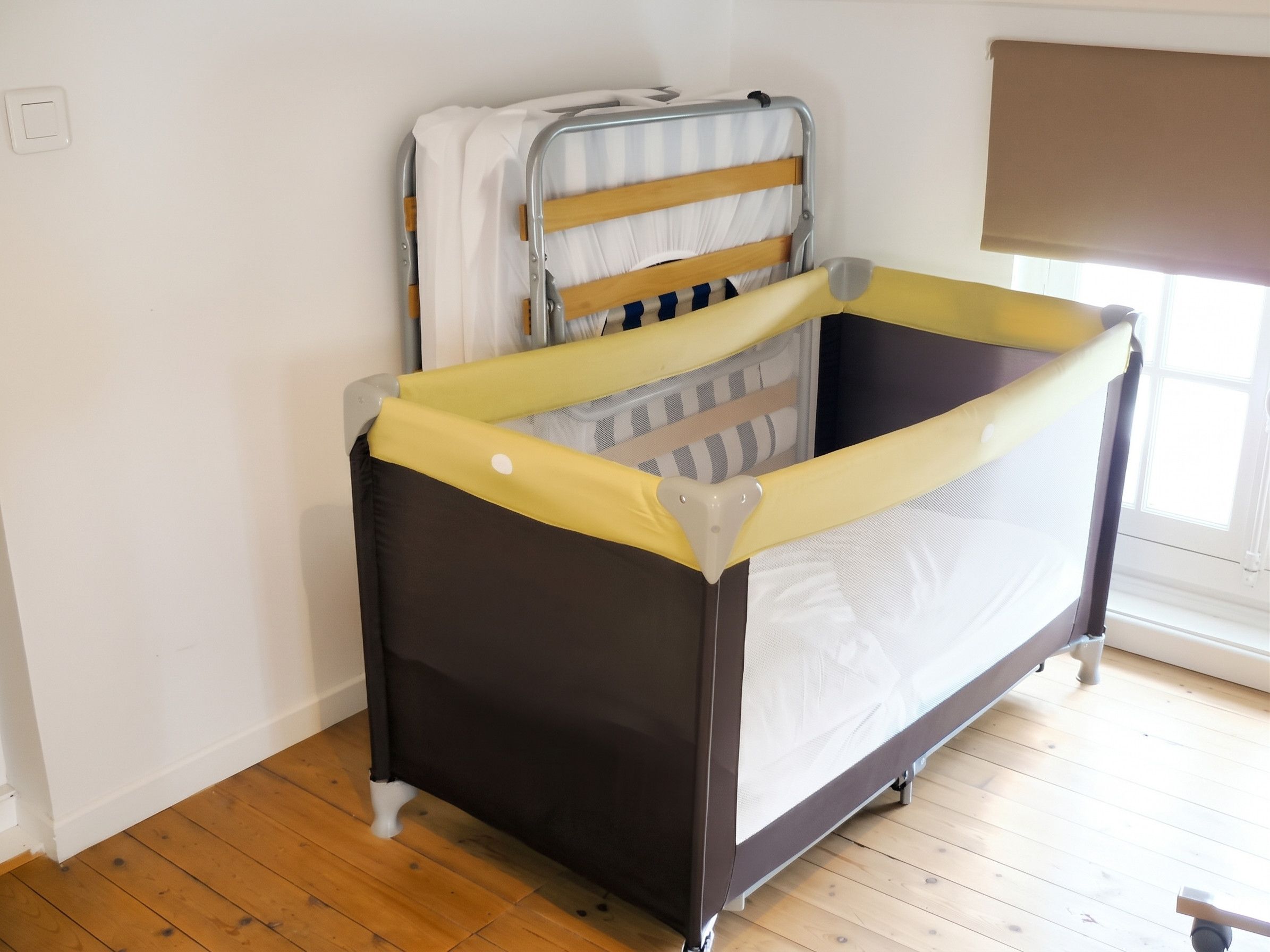 Un berceau pour bébé avec une barrière de sécurité jaune, un matelas recouvert d’une housse à rayures et une fenêtre dont le store est partiellement baissé.
