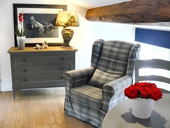 A room with a plaid armchair, a dresser with a mirror, a vase of red flowers, and a wooden beam above.