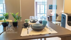 Una mesa de comedor dispuesta con un bol, platos, tazas y cubiertos, con plantas en macetas y vista de la habitación.