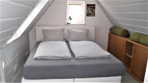 A small, narrow room with a sloped ceiling, a bed with white bedding, a small window with a desk lamp, and a wooden cabinet with green cushions.