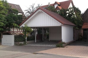 En etagers bygning med en carport, en murstenssti der fører til indgangen, og et lille haveareal med planter og blomster.