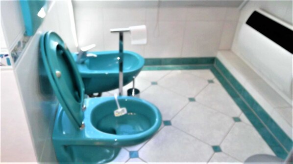 Baño pequeño con dos inodoros, un secador de manos y piso de baldosas.