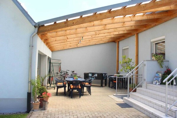 An outdoor patio area with a wooden pergola, a dining table with chairs, a grill, and potted plants.
