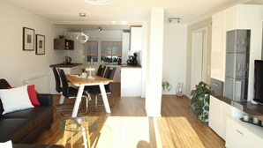Ein modernes Wohnzimmer mit einer Couch, einem Couchtisch und einem Essbereich mit Stühlen und einem Tisch. Im Hintergrund befindet sich eine Küche mit einem Kühlschrank und einer Mikrowelle.