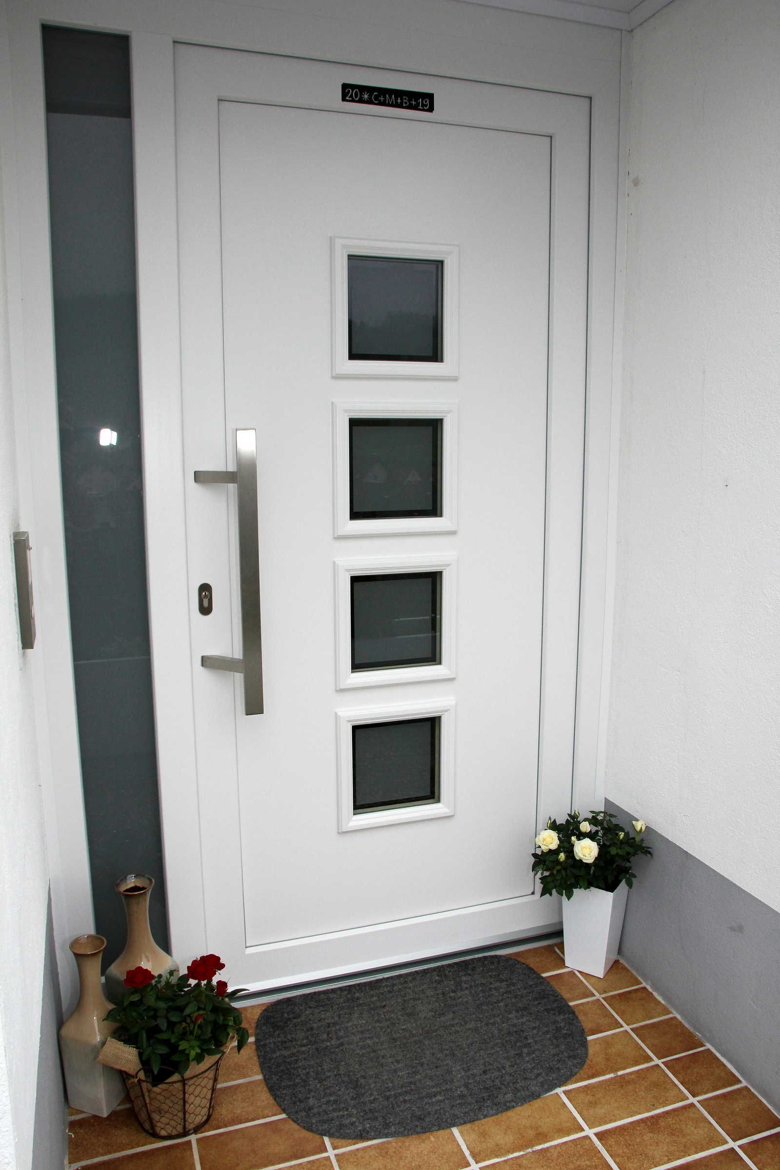 A white door with a handle and three windows, a doormat, and two potted plants with flowers.
