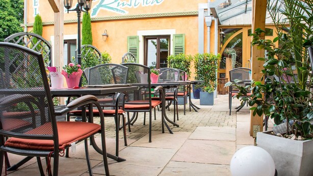 Un espace extérieur aménagé en terrasse, avec des chaises et des tables en métal, des plantes en pot et un bâtiment sur lequel est apposée une enseigne indiquant « Rustica ».