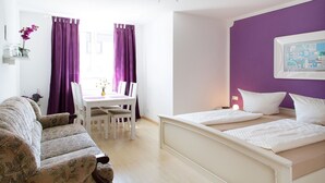 A room with a bed, a sofa, a dining table with chairs, and purple curtains. - Standard family room - Pension Sieg (Rust)