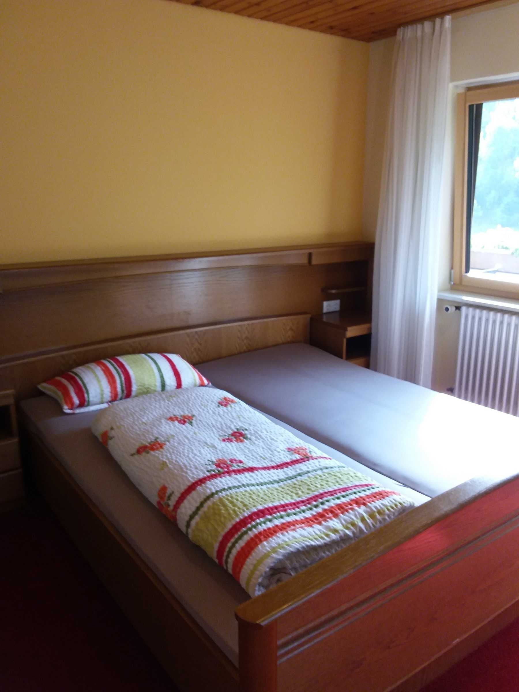 A single bed with a wooden headboard, a plain wall, and a window with curtains.